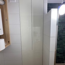 Meble na wymiar Bartosz Dopierała - Biała szafa łazienkowa z połyskiem, zintegrowana z zabudową, widoczna na tle kafelków. Otwarta szafka z drewnianym fornirem po lewej stronie.