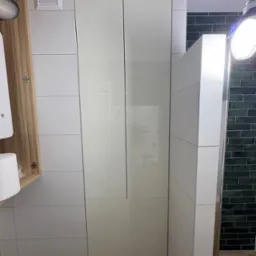 Biała szafa łazienkowa z połyskiem, zintegrowana z zabudową, widoczna na tle kafelków. Otwarta szafka z drewnianym fornirem po lewej stronie.