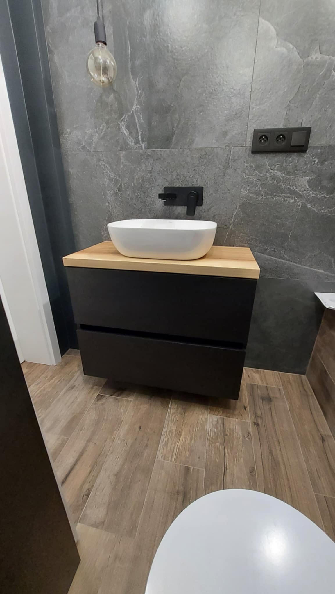 Nowoczesna łazienka z umywalką nablatową na szafce z czarnymi frontami i drewnianym blatem, szara ściana z marmurowym wzorem, oświetlenie żarówką Edisona.