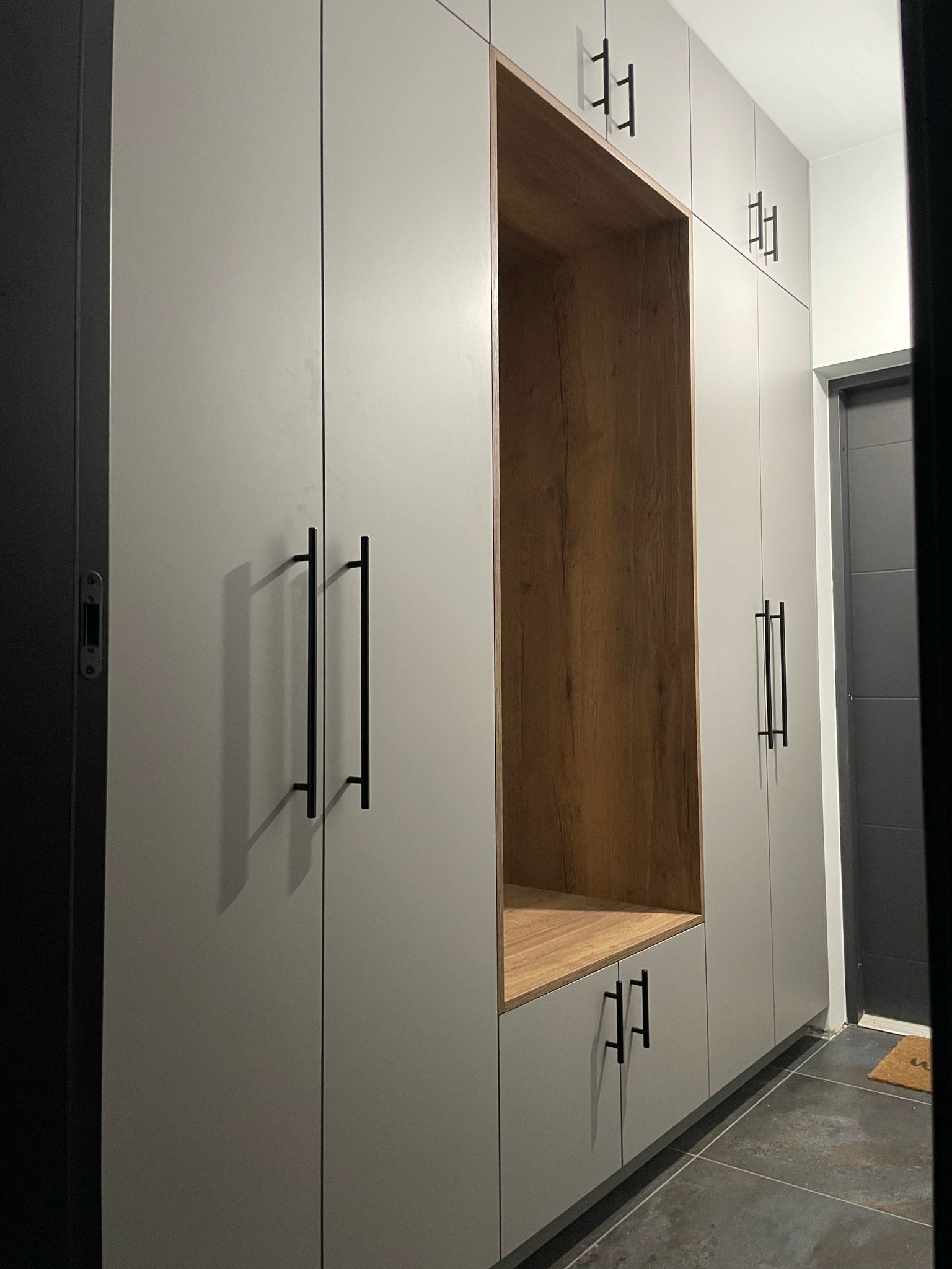 Szara zabudowa meblowa z drewnianymi akcentami, czarne uchwyty, widok na przedpokój z szarą podłogą i drzwiami.