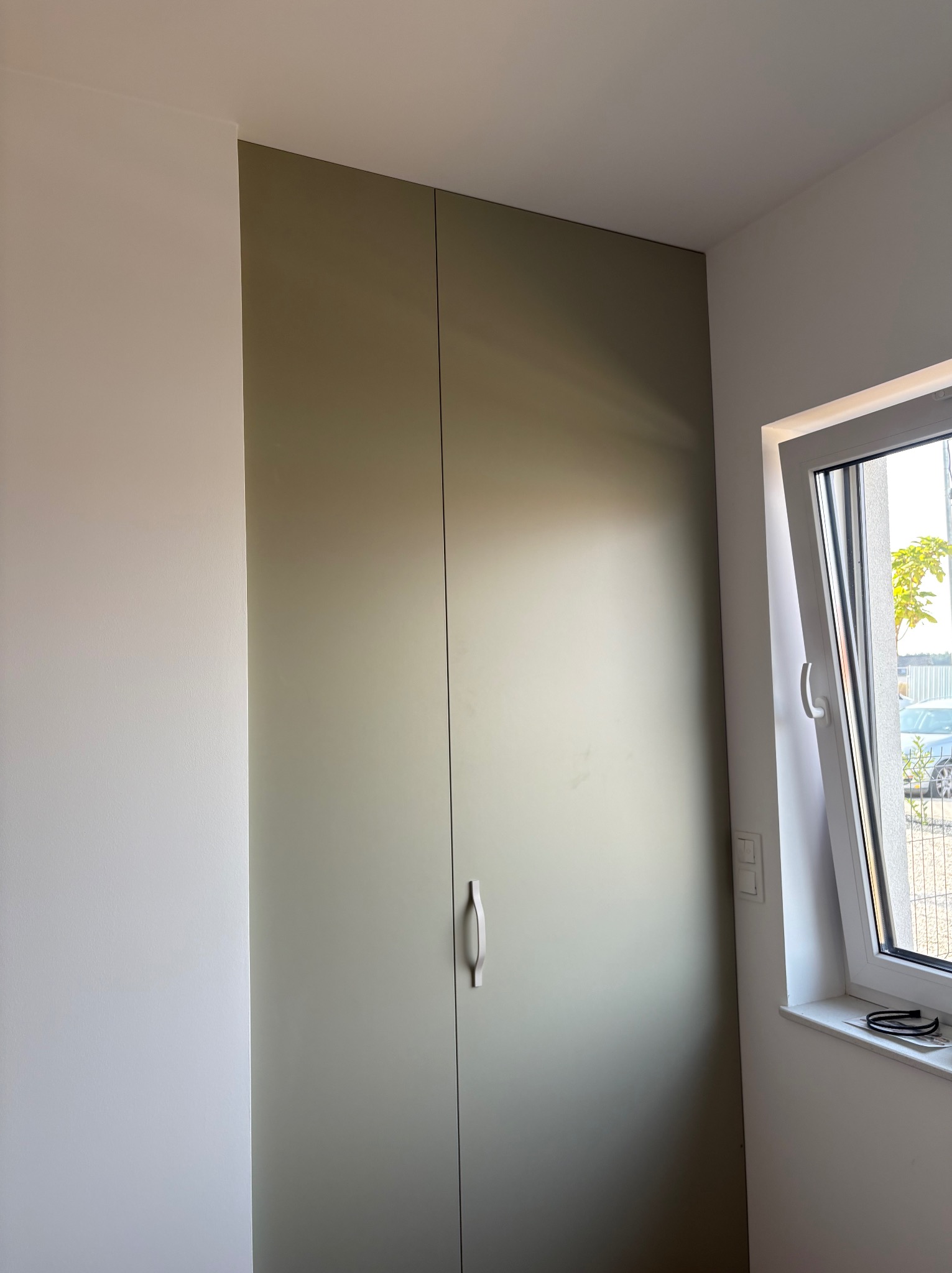 Wysoka szafa wnękowa z oliwkowymi drzwiami, z minimalistycznym uchwytem, w jasnym wnętrzu obok okna. Widok z dołu, podkreślający wysokość zabudowy.