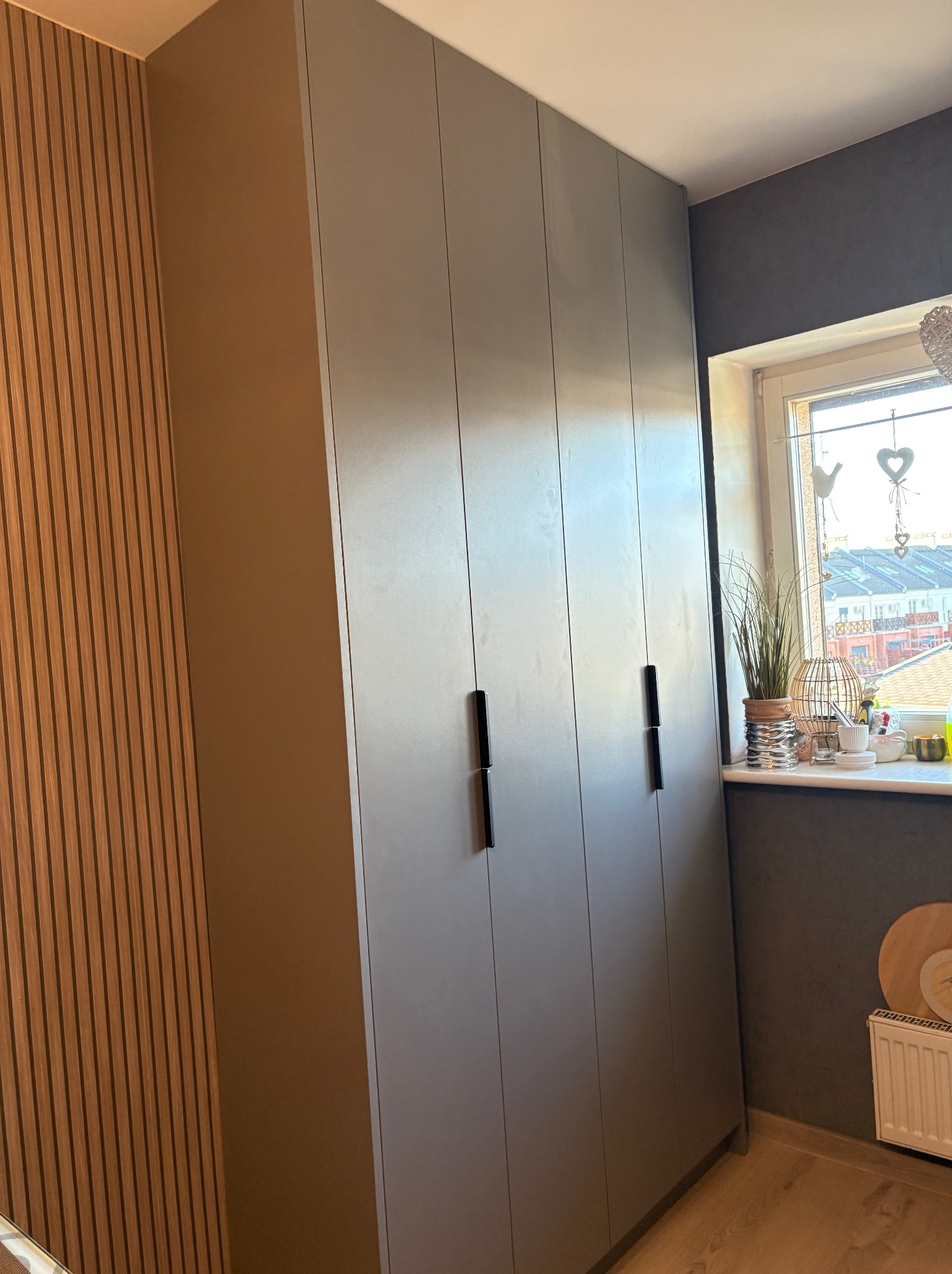Szara szafa wnękowa z czarnymi uchwytami, dopasowana do ściany obok okna, z dekoracyjnym panelem ściennym z drewnianych listewek po lewej stronie.