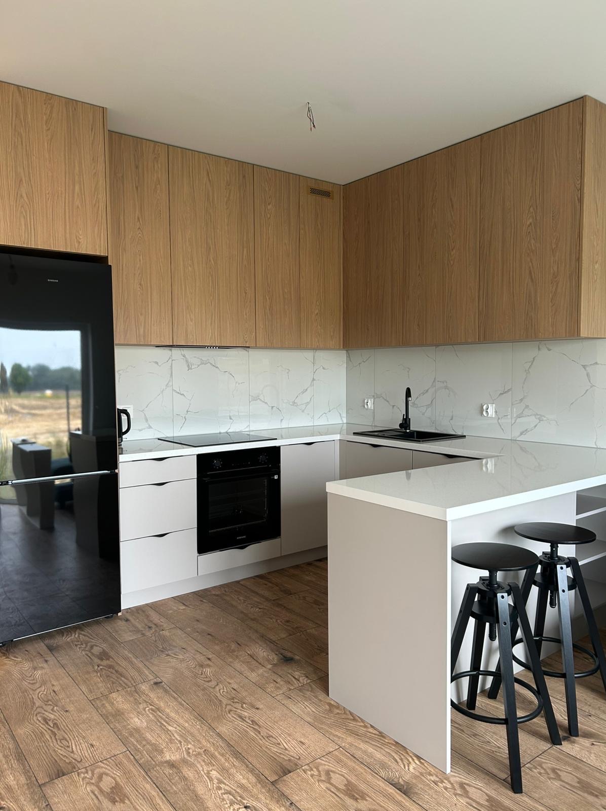 Nowoczesna kuchnia z drewnianymi szafkami górnymi i białymi dolnymi. Czarne krzesła barowe przy wyspie. Podłoga drewniana. Widok na ogród z okna.