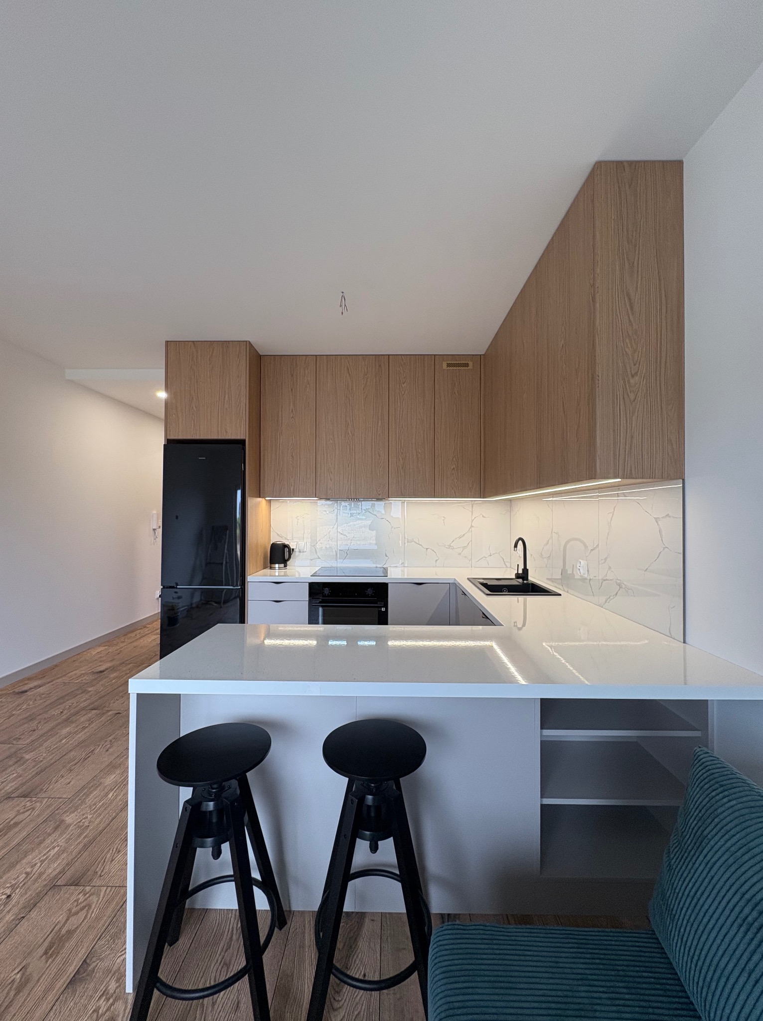 Nowoczesna kuchnia z drewnianymi szafkami, marmurowym blatem i czarnymi detalami: lodówka, bateria i hokery. Oświetlenie LED podkreśla elegancję.