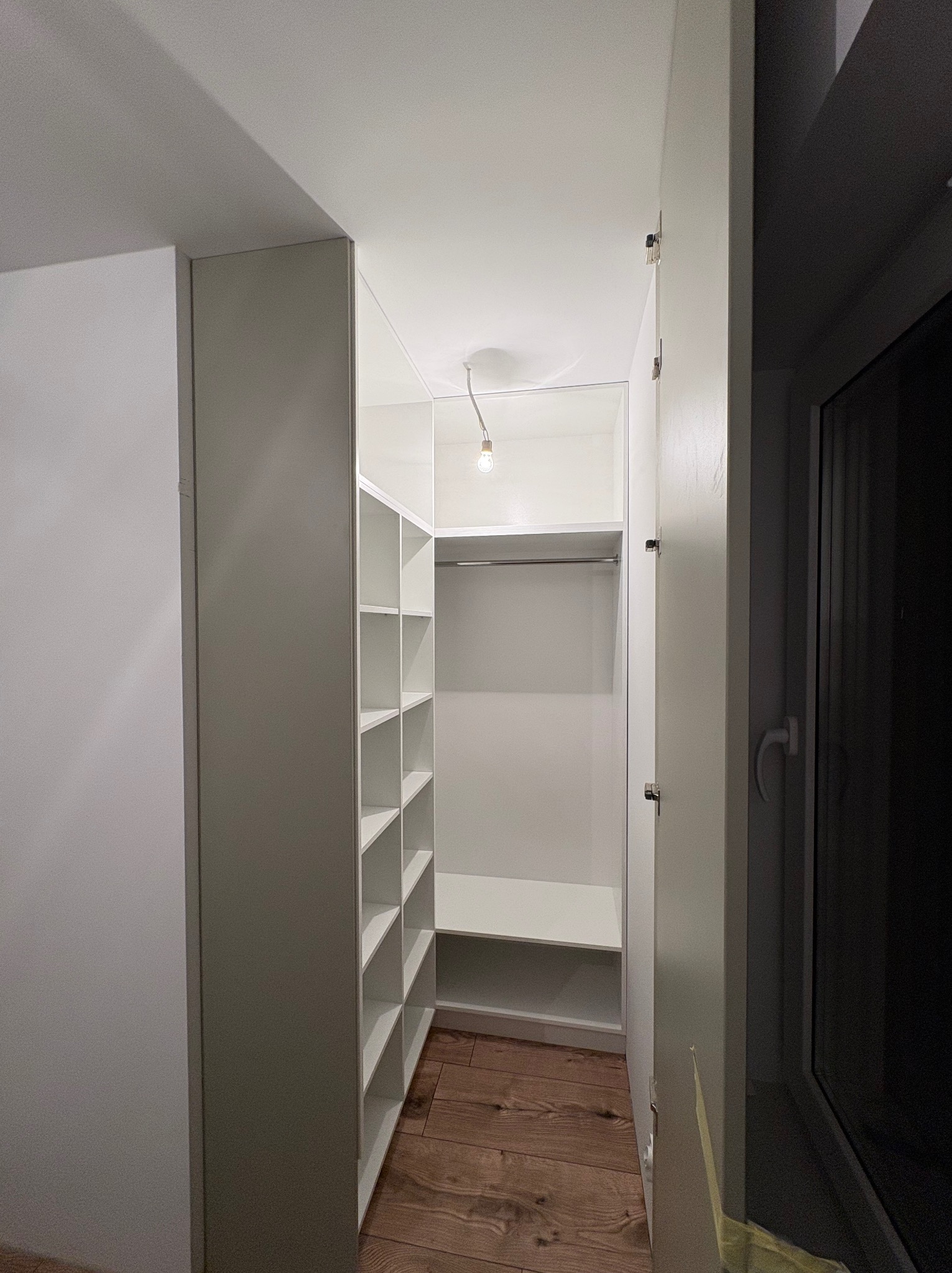 Biała szafa wnękowa z półkami i drążkiem na ubrania, widok z wnętrza, drewniana podłoga, minimalistyczny styl, oświetlenie żarówką.