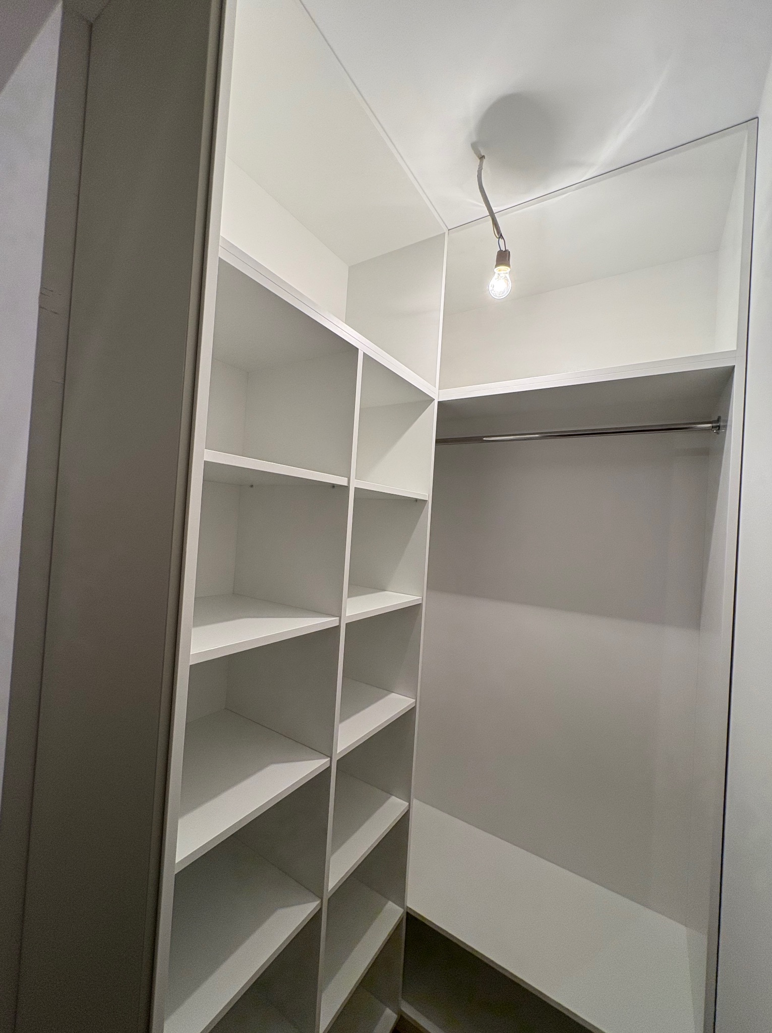 Wnętrze szafy wnękowej z półkami po lewej i drążkiem na ubrania po prawej stronie. Białe wykończenie, oświetlenie punktowe u góry.