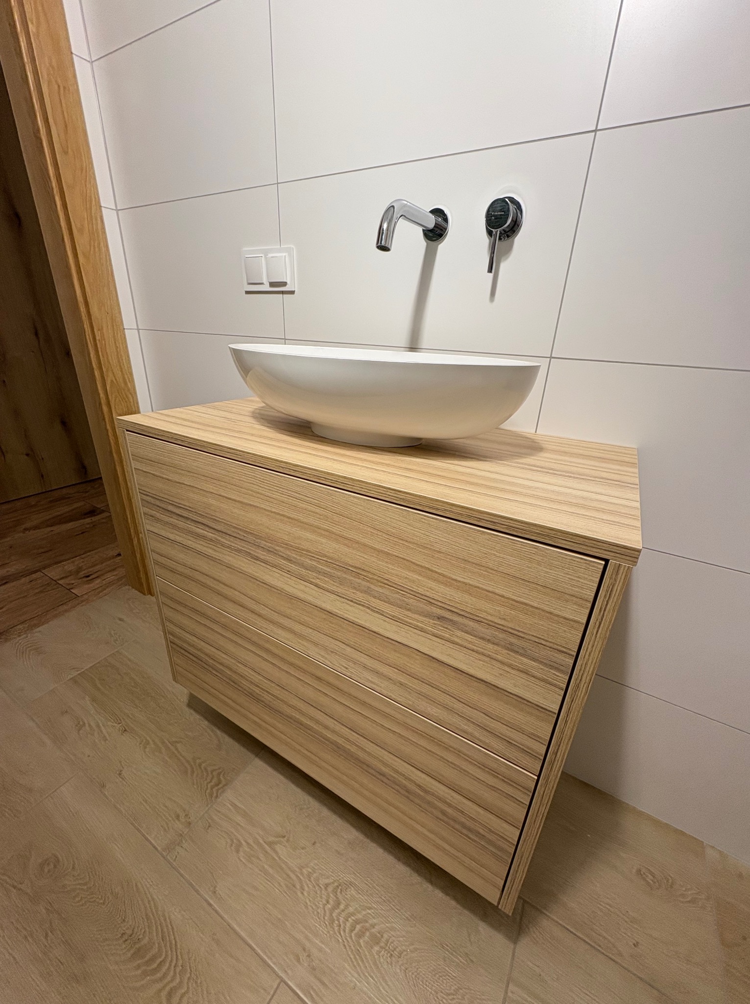 Nowoczesna łazienka: drewniana szafka pod umywalkę nablatową z chromowaną armaturą na tle białych płytek. Minimalistyczny design, ciepłe oświetlenie.