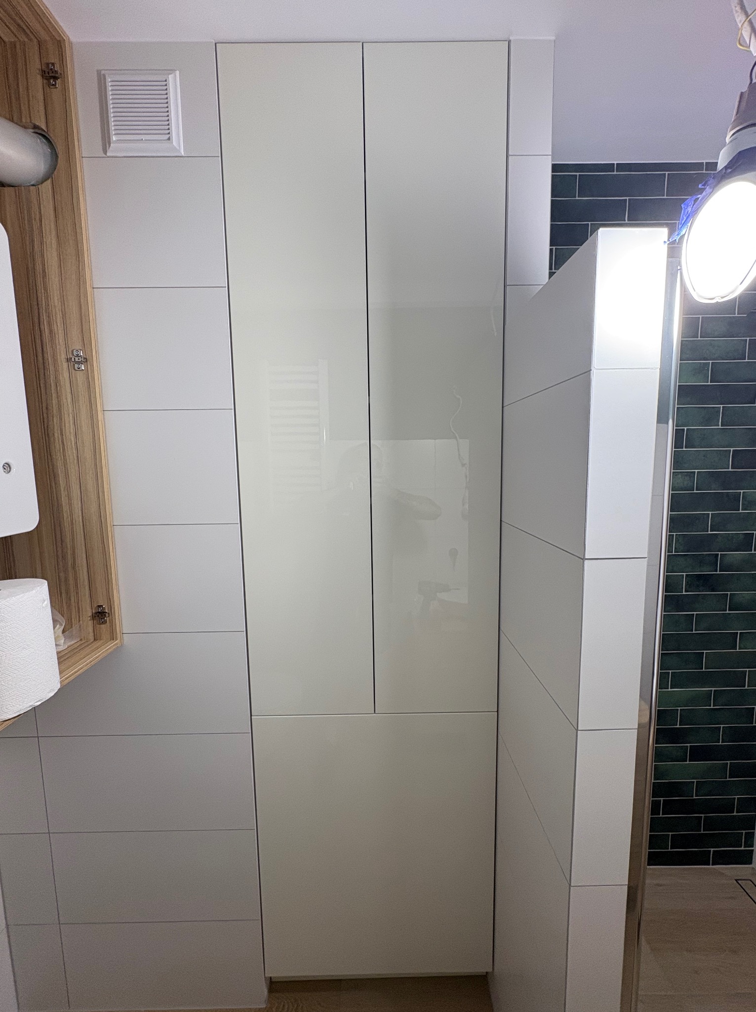 Biała szafa łazienkowa z połyskiem, zintegrowana z zabudową, widoczna na tle kafelków. Otwarta szafka z drewnianym fornirem po lewej stronie.
