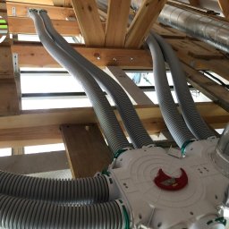 ECOrekuperacje Maciej Leśniewicz - Rozdzielacz powietrza z podłączonymi elastycznymi przewodami wentylacyjnymi w surowym wnętrzu poddasza, widoczne elementy drewnianej konstrukcji dachu.