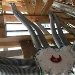 Rozdzielacz powietrza z podłączonymi elastycznymi przewodami wentylacyjnymi w surowym wnętrzu poddasza, widoczne elementy drewnianej konstrukcji dachu.