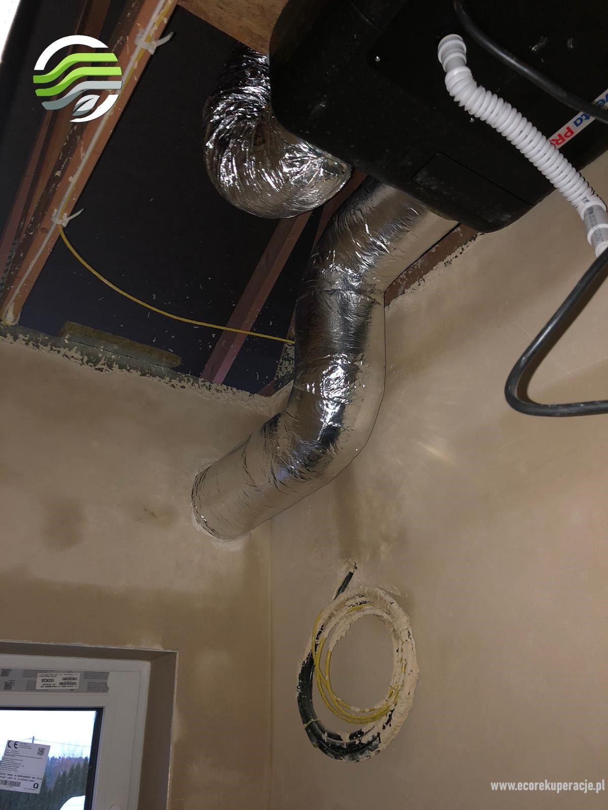 Instalacja systemu wentylacyjnego z rurami aluminiowymi w nowym budynku, widoczne przewody elektryczne, okno i fragment sufitu.