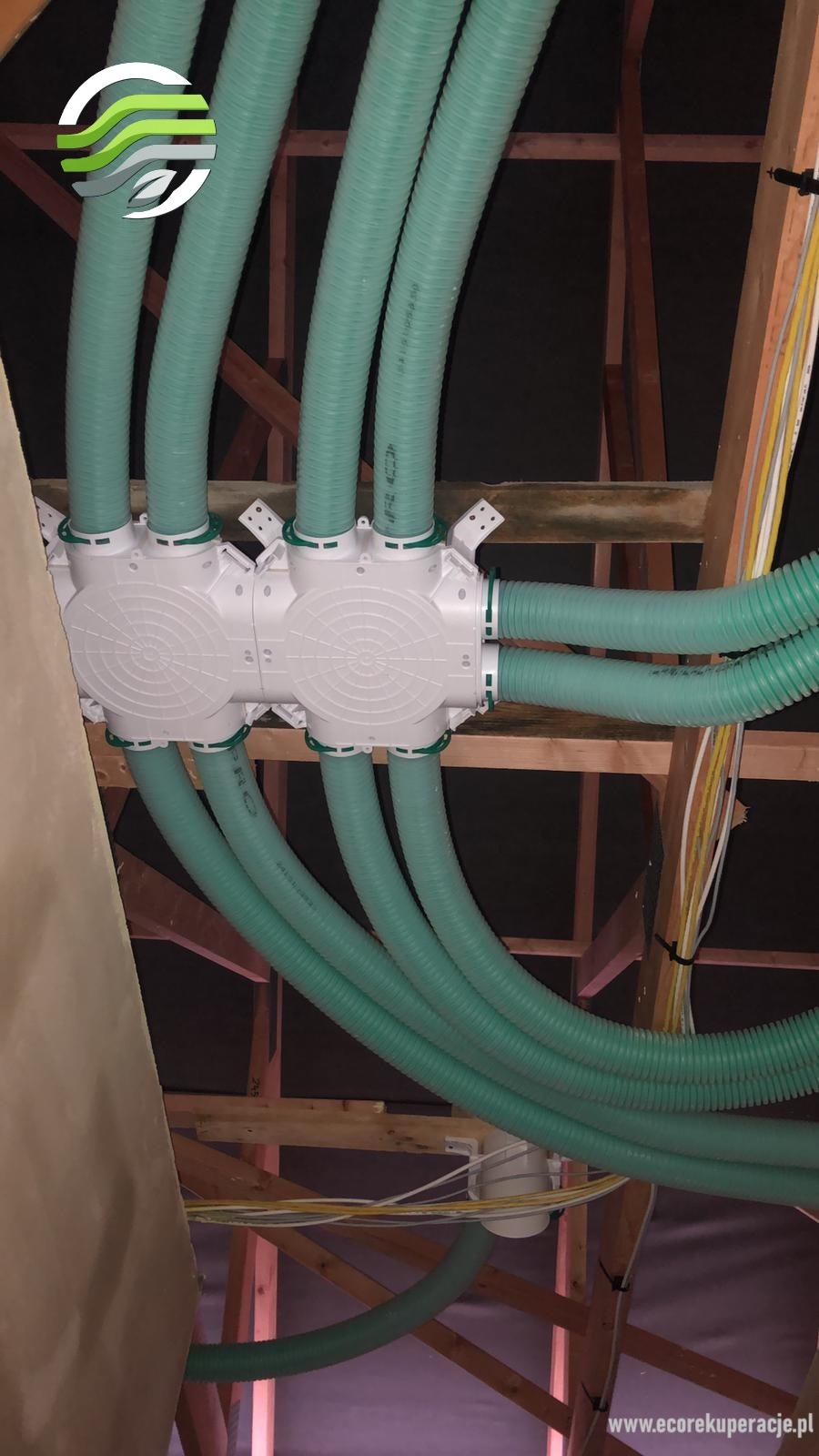 Rozdzielacz wentylacyjny z podłączonymi elastycznymi przewodami wentylacyjnymi w trakcie montażu na poddaszu, widoczne elementy drewnianej konstrukcji dachu.