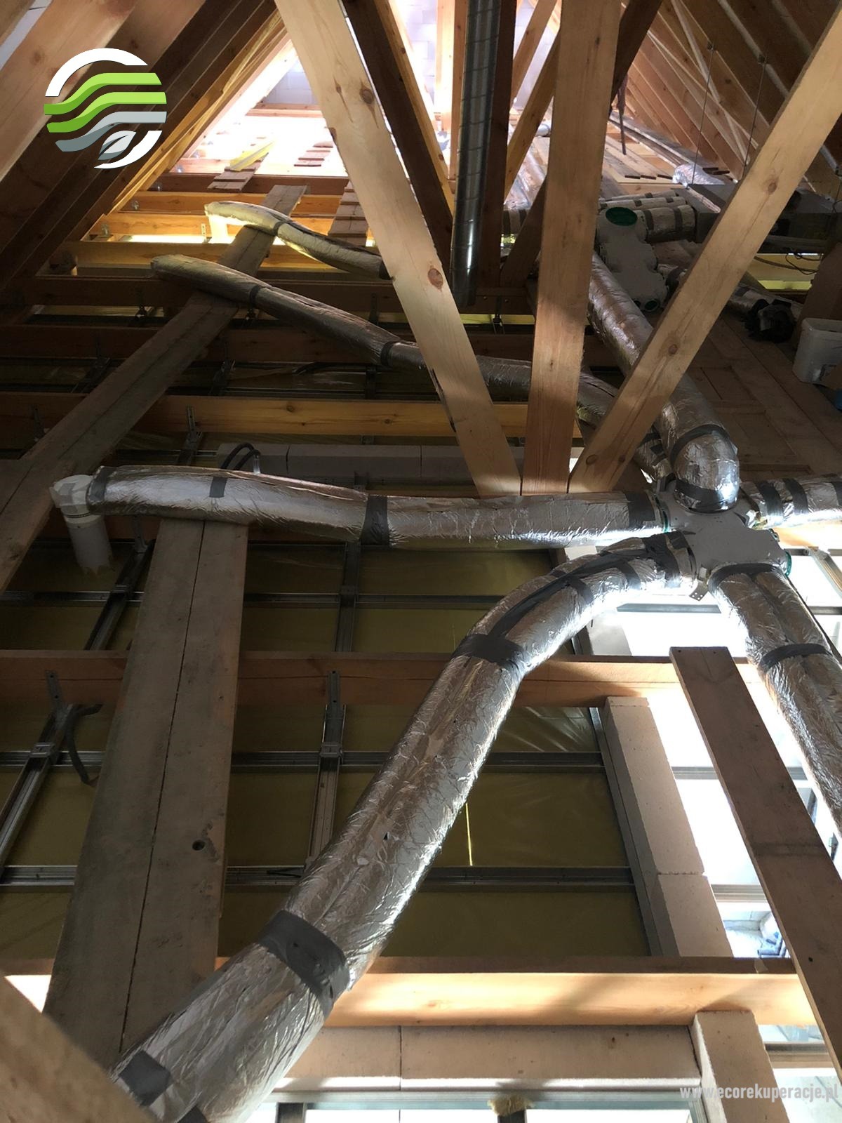Widok z dołu na system kanałów wentylacyjnych w folii aluminiowej, zamontowany w przestrzeni poddasza z drewnianą konstrukcją więźby dachowej i żółtą izolacją.