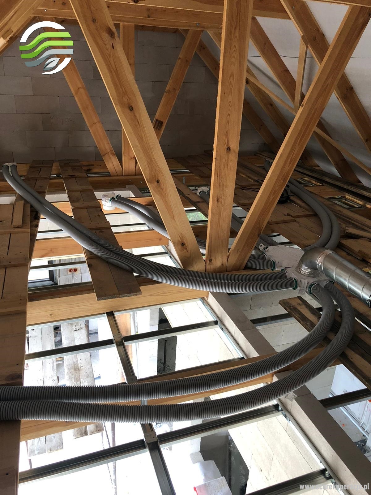 Szare, karbowane przewody wentylacyjne rozprowadzone w przestrzeni poddasza z drewnianą konstrukcją więźby dachowej i oknami dachowymi w tle.