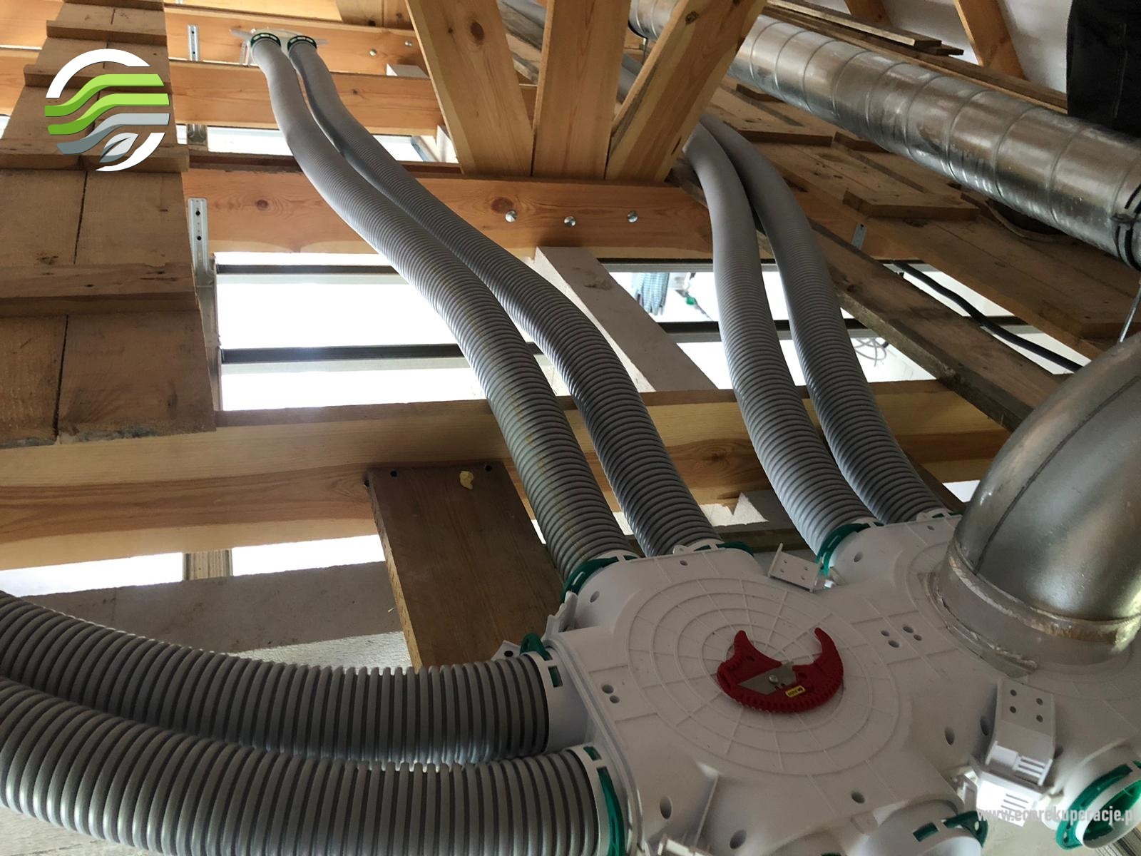 Rozdzielacz powietrza z podłączonymi elastycznymi przewodami wentylacyjnymi w surowym wnętrzu poddasza, widoczne elementy drewnianej konstrukcji dachu.