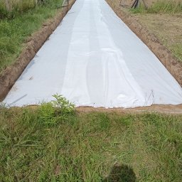 Kop-tech wladyslaw kawecki - Szeroki wykop z rozłożoną białą geowłókniną, przygotowany pod dalsze prace ziemne. W tle widoczny koparka i hałda ziemi, co sugeruje trwające roboty.