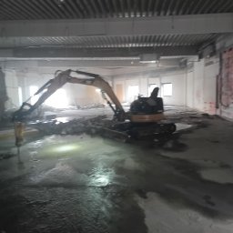 Kop-tech wladyslaw kawecki - Żółta koparka z hydraulicznym młotem pneumatycznym wewnątrz budynku w trakcie prac rozbiórkowych. Widoczne gruz i woda na podłodze, surowe ściany i sufit.