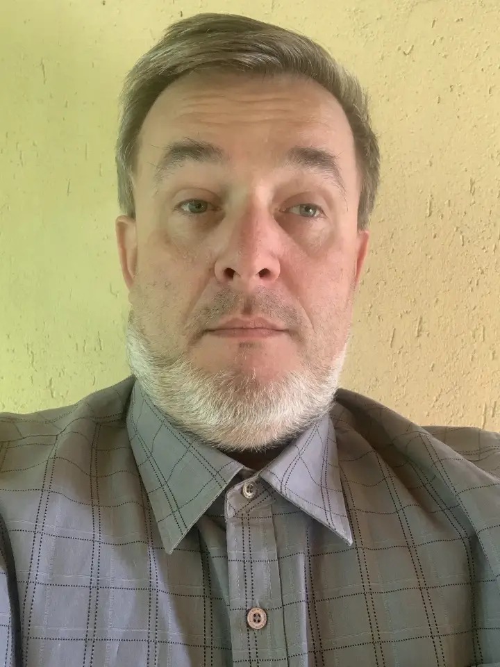 Portret mężczyzny w szarej koszuli w kratę na tle żółtej ściany. Widoczne zmarszczki, szpakowata broda. Ujęcie od pasa w górę, naturalne światło.