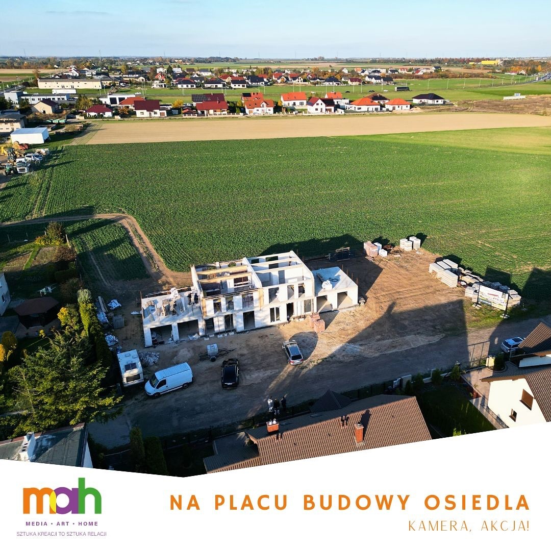 Widok z lotu ptaka na plac budowy osiedla domów jednorodzinnych w stanie surowym otwartym, z widocznymi materiałami budowlanymi i zaparkowanymi pojazdami na tle zielonego pola i zabudowań...