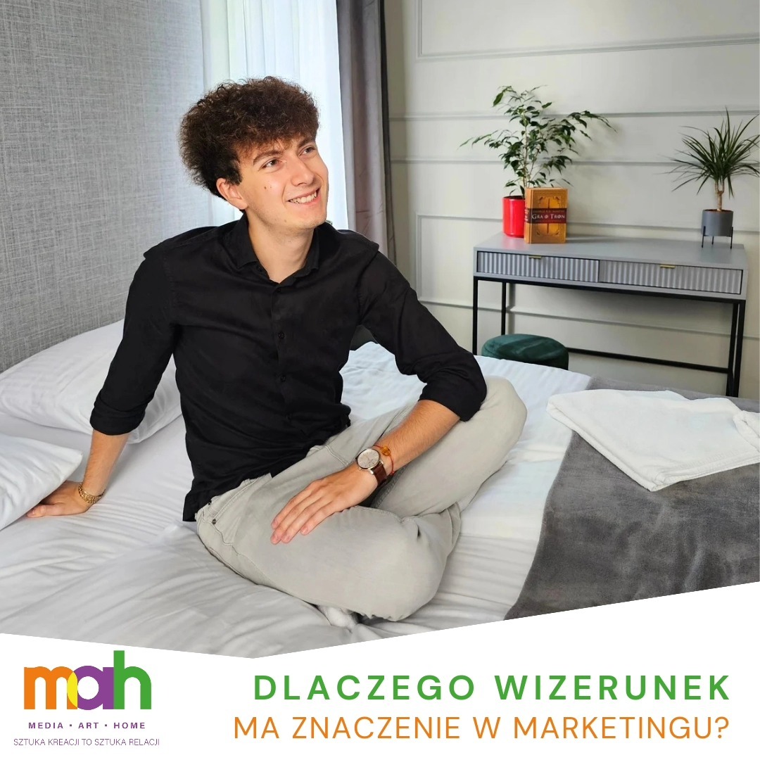 Młody mężczyzna z kręconymi włosami siedzi na łóżku w czarnej koszuli i jasnych spodniach, uśmiecha się i patrzy w bok. W tle widoczny stolik z roślinami. U dołu grafika z pytaniem: 'Dlaczego...