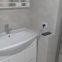 Łazienka z umywalką nablatową, białą szafką pod umywalką, baterią umywalkową i beżowymi płytkami na ścianie. Na blacie obok umywalki leży ładujący się smartfon.