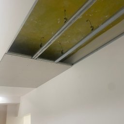 VIKTOR HERASYMIV - Montaż sufitu podwieszanego z płyt karton-gips na metalowej konstrukcji. Widoczne kable oświetleniowe. Świeżo zamontowana płyta. Białe ściany w tle.