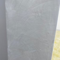 VIKTOR HERASYMIV - Ściana po nałożeniu szarej gładzi, widoczne ślady po szpachlowaniu. Fragment zabezpieczonej podłogi w rogu kadru. Delikatna faktura i neutralna kolorystyka.