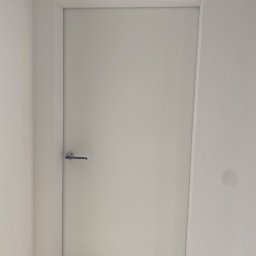 VIKTOR HERASYMIV - Białe, minimalistyczne drzwi wewnętrzne z srebrną klamką, wkomponowane w białą ścianę. Proste wykończenie, brak dekoracji, jednolite oświetlenie.