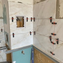 VIKTOR HERASYMIV - Układanie płytek imitujących marmur na ścianie z widocznymi systemami poziomowania. W tle instalacja wodna i fragment ściany z płyt karton-gips.