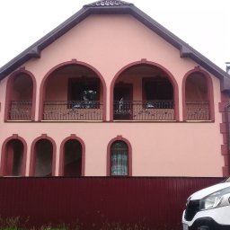 VIKTOR HERASYMIV - Dwukondygnacyjny dom z balkonem i łukowymi oknami, pomalowany na różowo z bordowymi akcentami. Widoczny fragment białego samochodu i drabina z boku budynku.