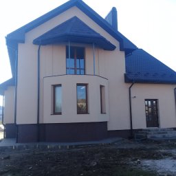 VIKTOR HERASYMIV - Elewacja domu jednorodzinnego w beżowej tonacji z ciemną podmurówką i dachem. Widoczne okna z drewnianymi ramami i wejście z betonowymi schodami. Teren wokół domu częściowo zagospodarowany.