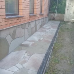 VIKTOR HERASYMIV - Wykończony fragment elewacji budynku z dekoracyjnym kamieniem i płytami chodnikowymi w odcieniach beżu i szarości, widok z zewnątrz, ceglana ściana z oknami w tle.