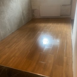 VIKTOR HERASYMIV - Ułożona podłoga z płytek imitujących drewno w pomieszczeniu z surowymi ścianami i grzejnikiem. Widoczny fragment czarnej, marmurkowej podłogi w przejściu.