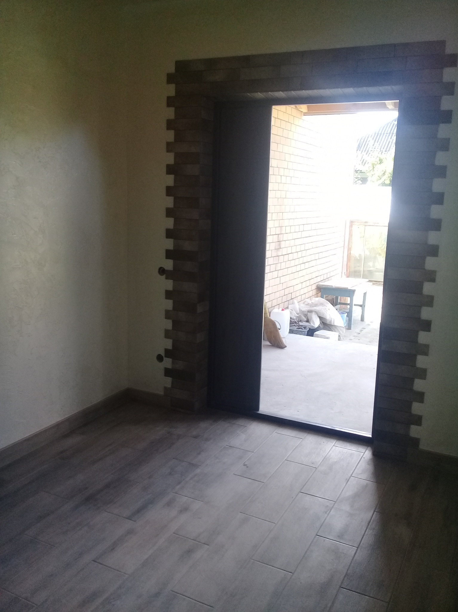 Wykończone wnętrze z brązową podłogą imitującą drewno i obramowaniem drzwi z cegiełek, widok na zewnątrz