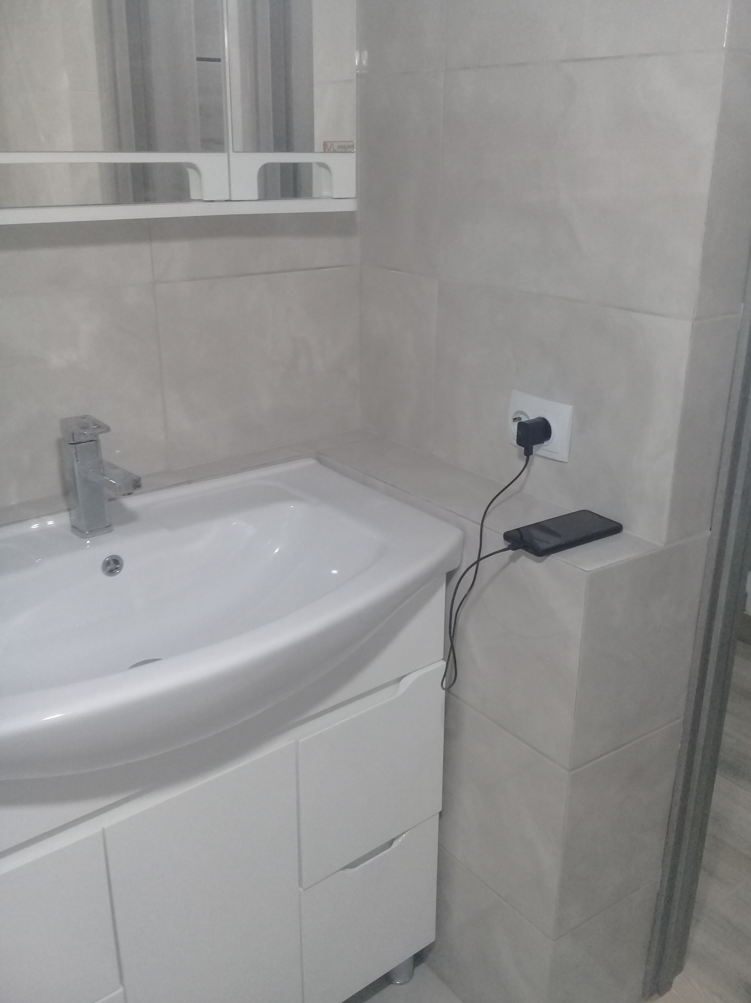 Łazienka z umywalką nablatową, białą szafką pod umywalką, baterią umywalkową i beżowymi płytkami na ścianie. Na blacie obok umywalki leży ładujący się smartfon.