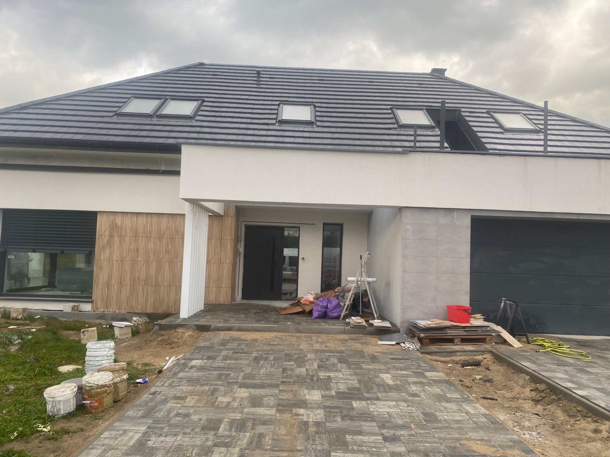 Nowoczesny dom jednorodzinny z szarą dachówką i kostką brukową na podjeździe. Widoczne detale wykończeniowe i elementy placu budowy.