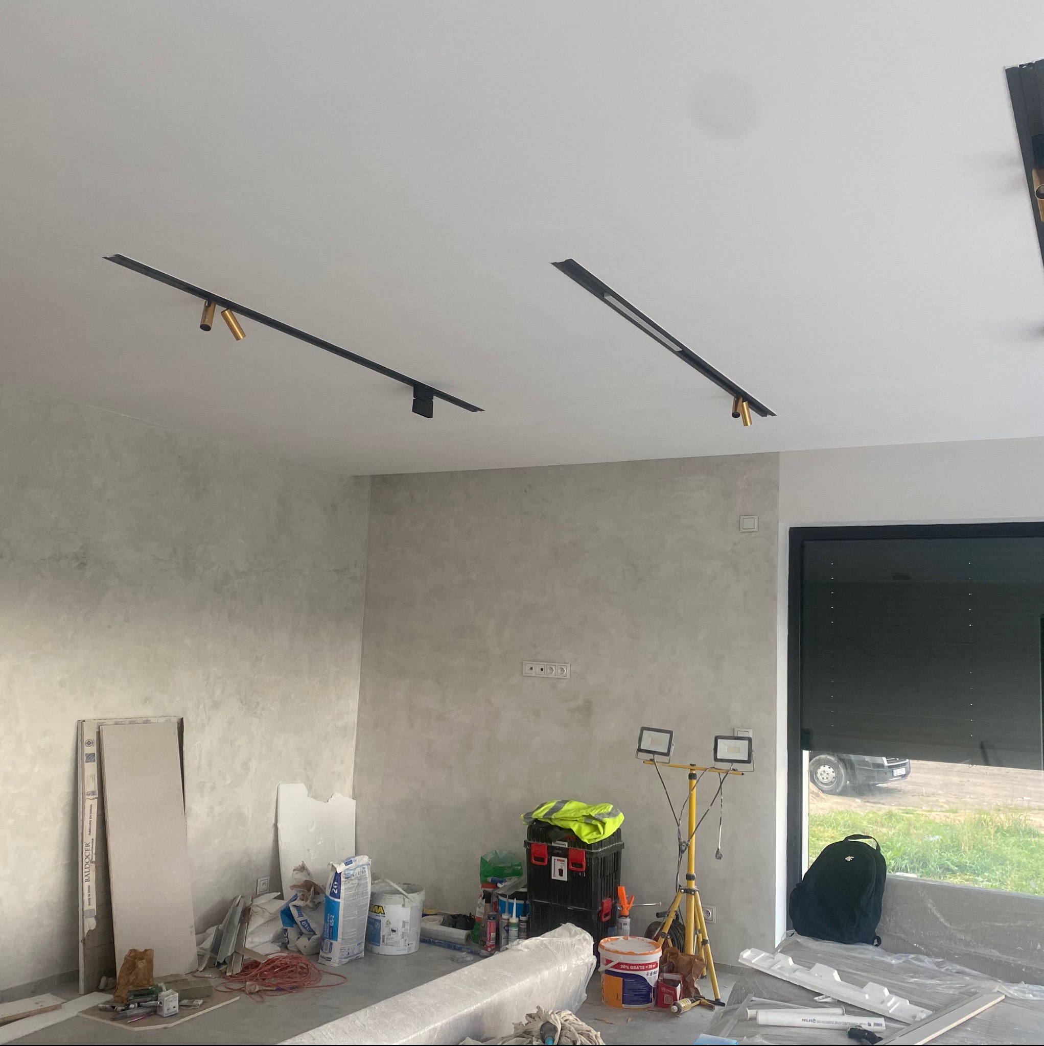 Pokój w trakcie wykańczania: ściany pokryte dekoracyjną szarą farbą, zamontowane czarne szyny oświetleniowe z reflektorami. Widoczne narzędzia i materiały budowlane.
