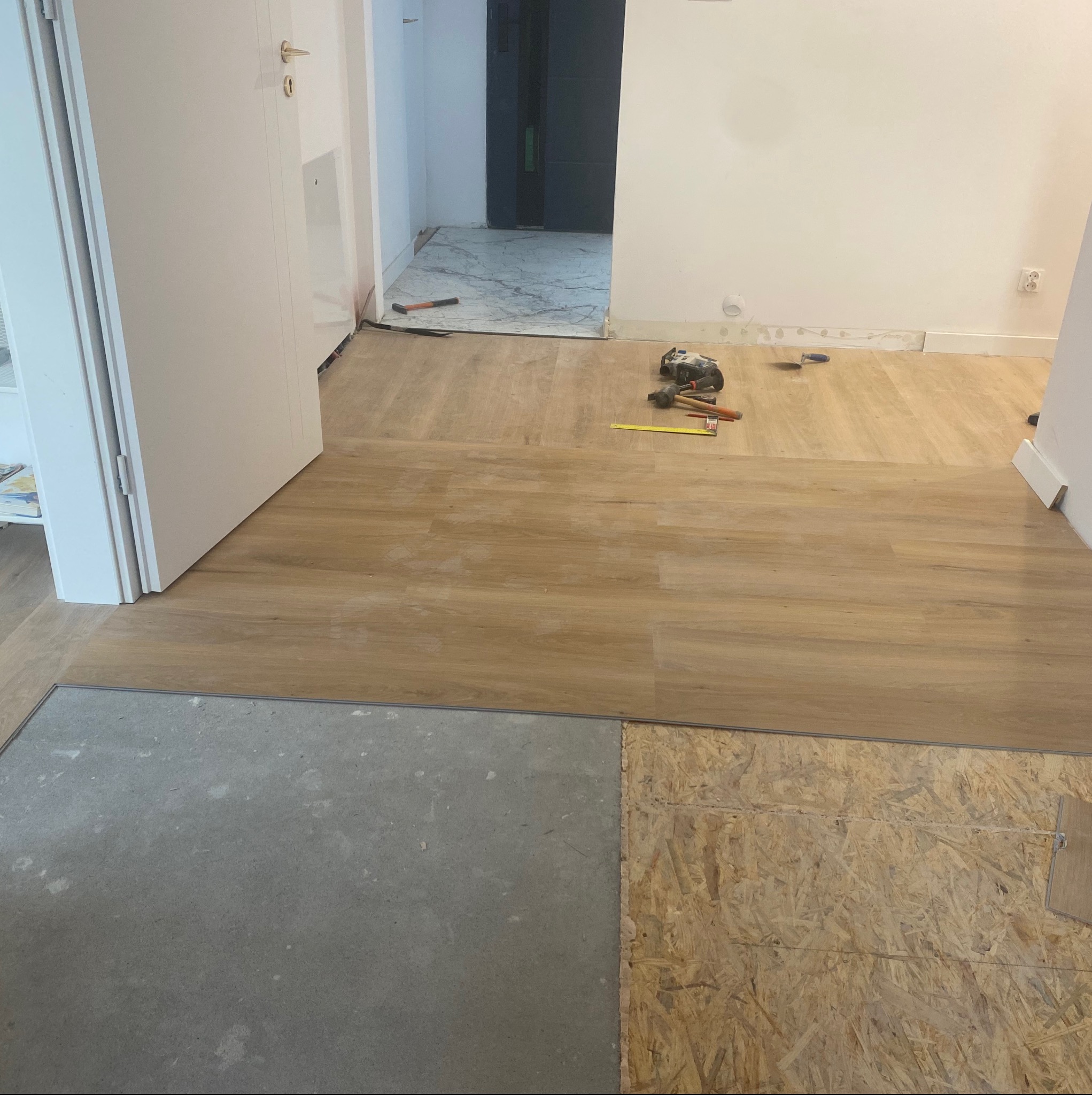 Układanie paneli podłogowych w trakcie remontu. Widoczne narzędzia, fragmenty podłoża (beton, płyta OSB) i ułożona część paneli w odcieniach jasnego drewna.