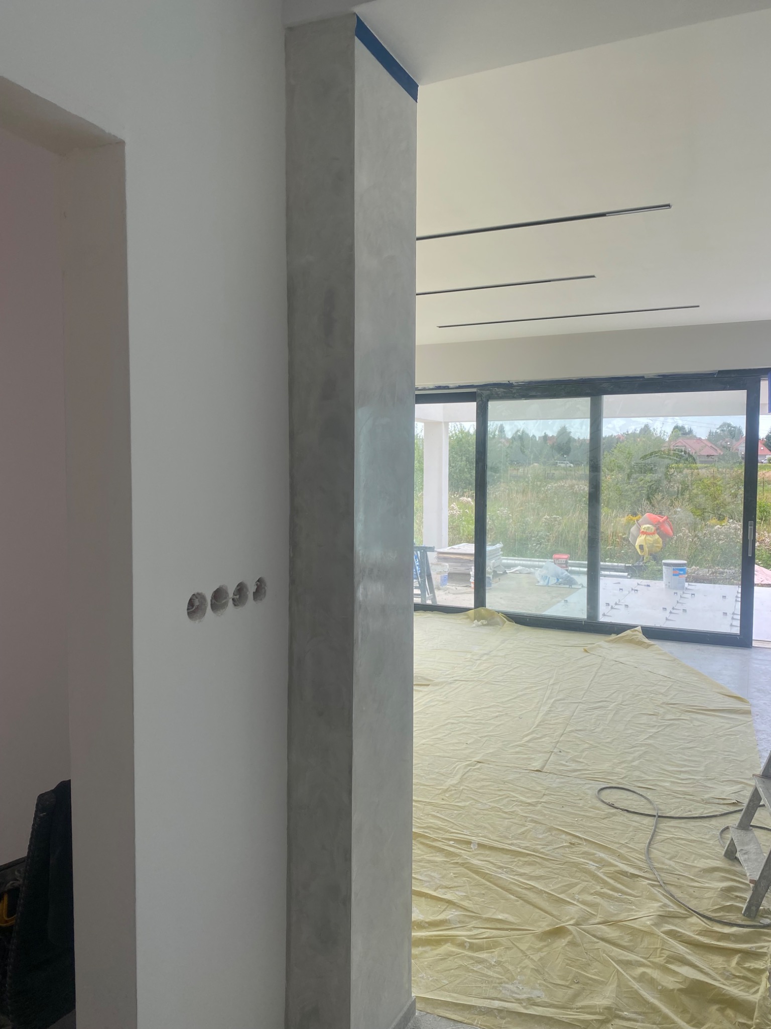 Wnętrze w trakcie wykańczania: kolumna z efektem betonu, ściana z gniazdkami, podłoga zabezpieczona folią malarską, okno przesuwne z widokiem na zewnątrz.