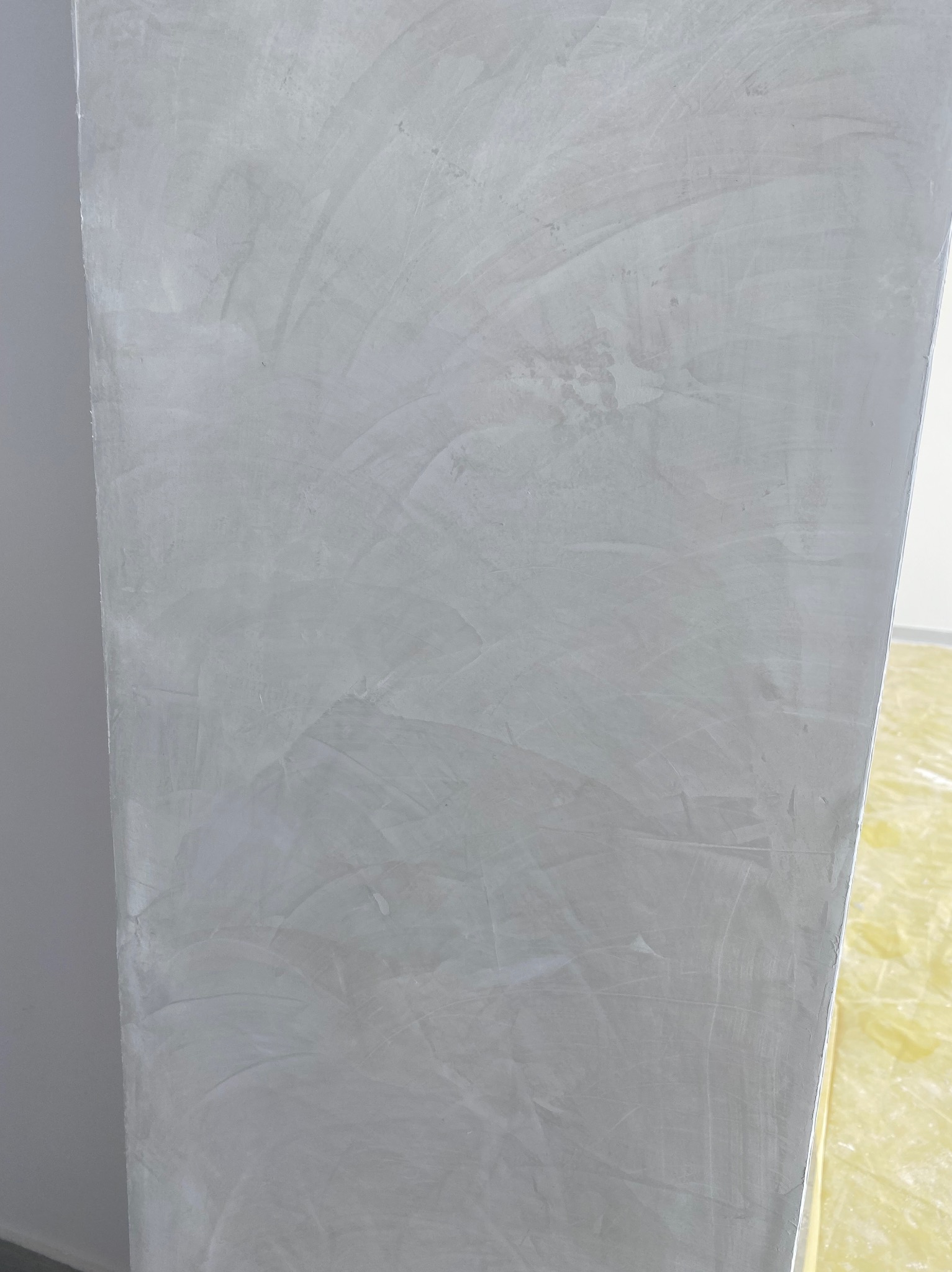 Ściana po nałożeniu szarej gładzi, widoczne ślady po szpachlowaniu. Fragment zabezpieczonej podłogi w rogu kadru. Delikatna faktura i neutralna kolorystyka.