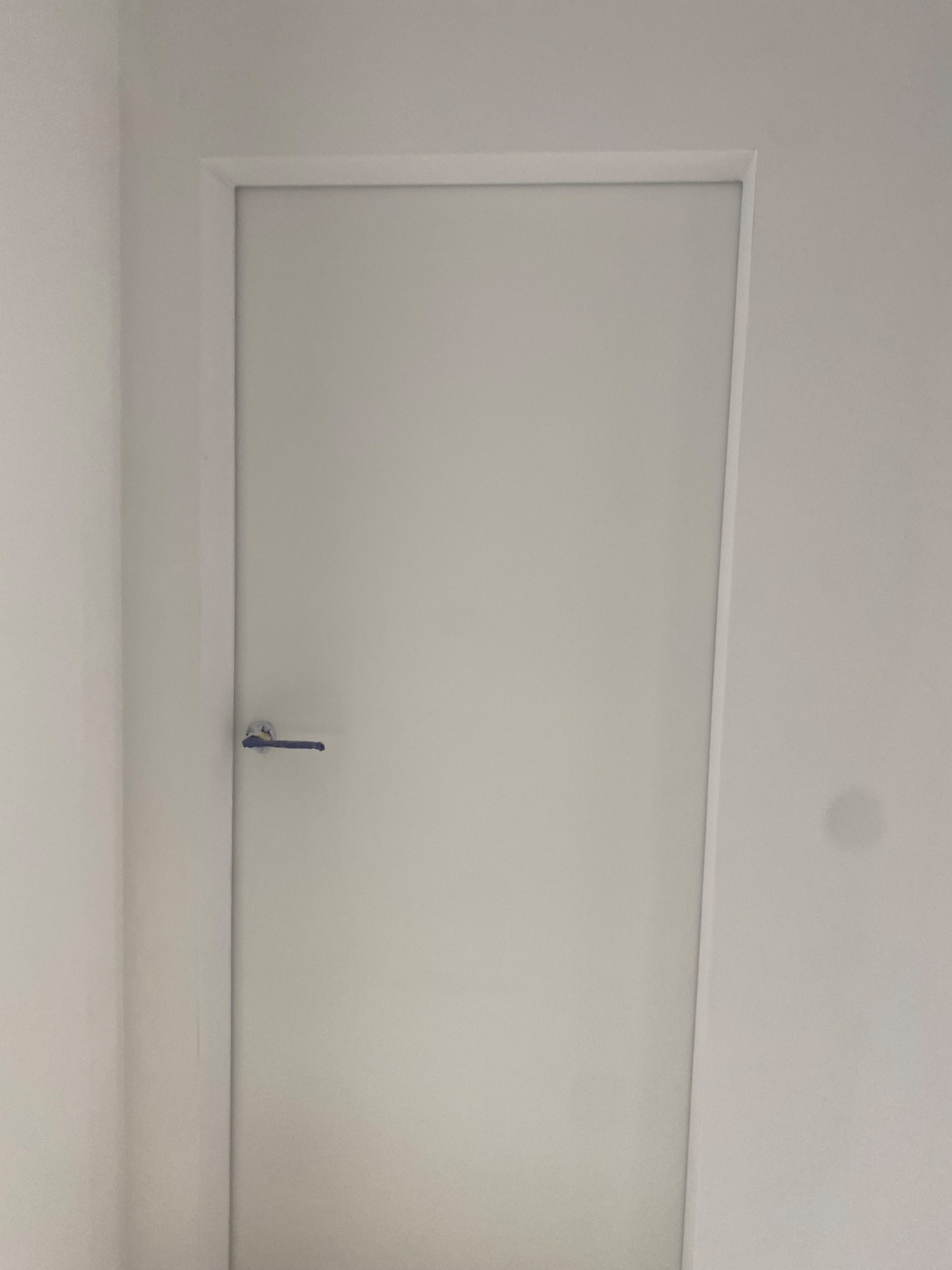 Białe, minimalistyczne drzwi wewnętrzne z srebrną klamką, wkomponowane w białą ścianę. Proste wykończenie, brak dekoracji, jednolite oświetlenie.