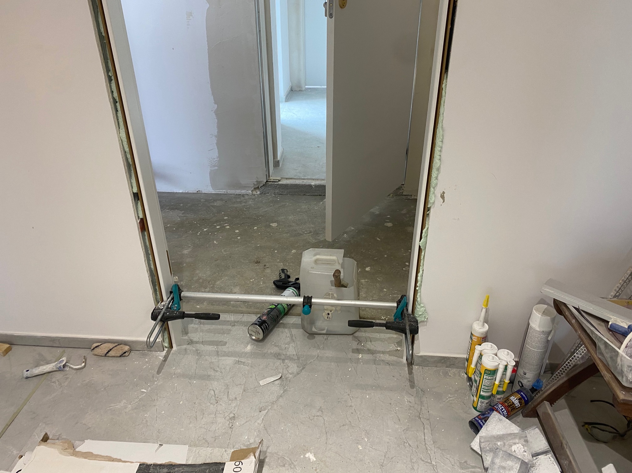 Montaż ościeżnicy drzwiowej z użyciem rozpórki i pianki montażowej. Widoczne narzędzia i materiały budowlane. W tle wykończone ściany i podłoga.