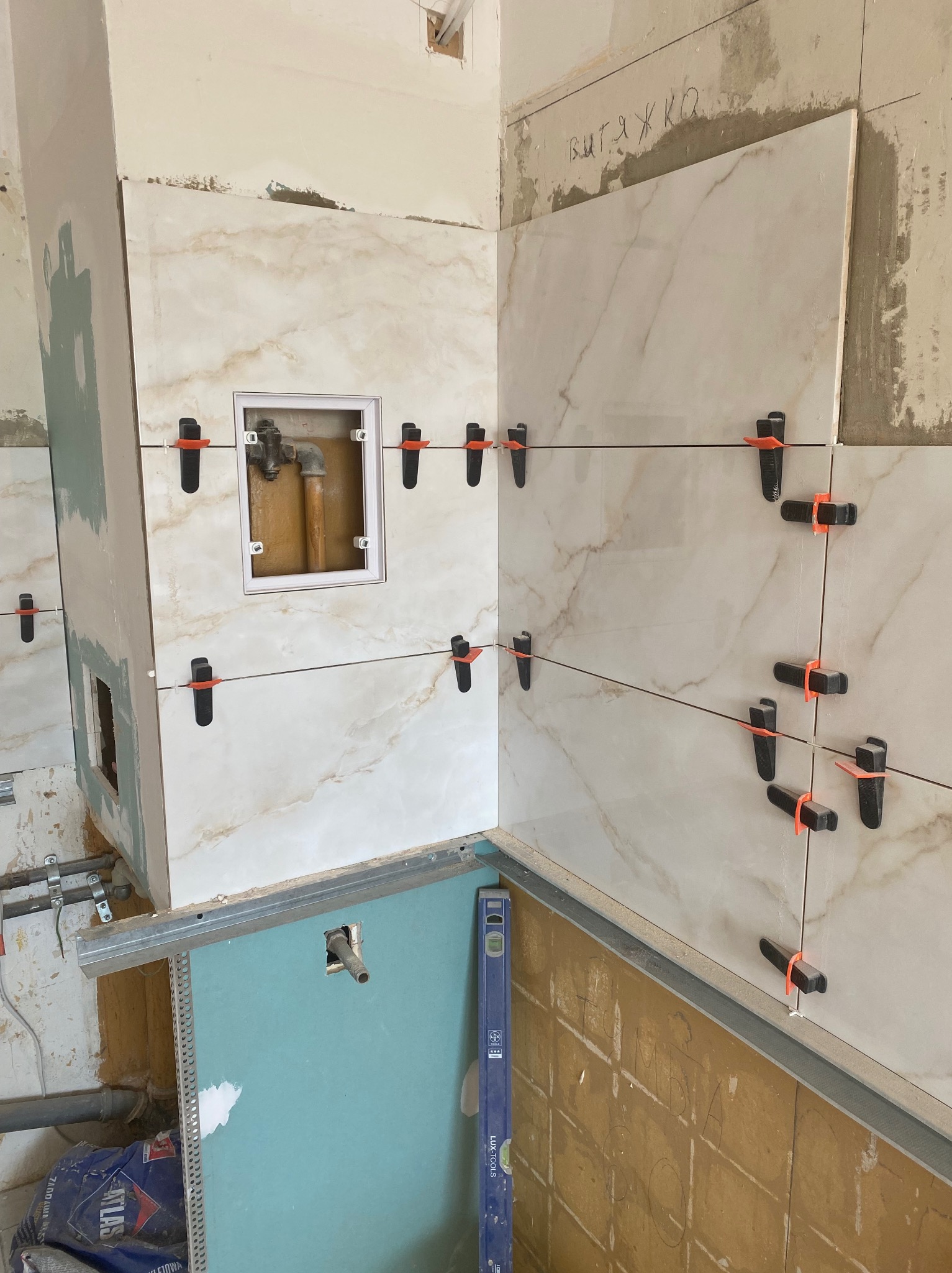 Układanie płytek imitujących marmur na ścianie z widocznymi systemami poziomowania. W tle instalacja wodna i fragment ściany z płyt karton-gips.