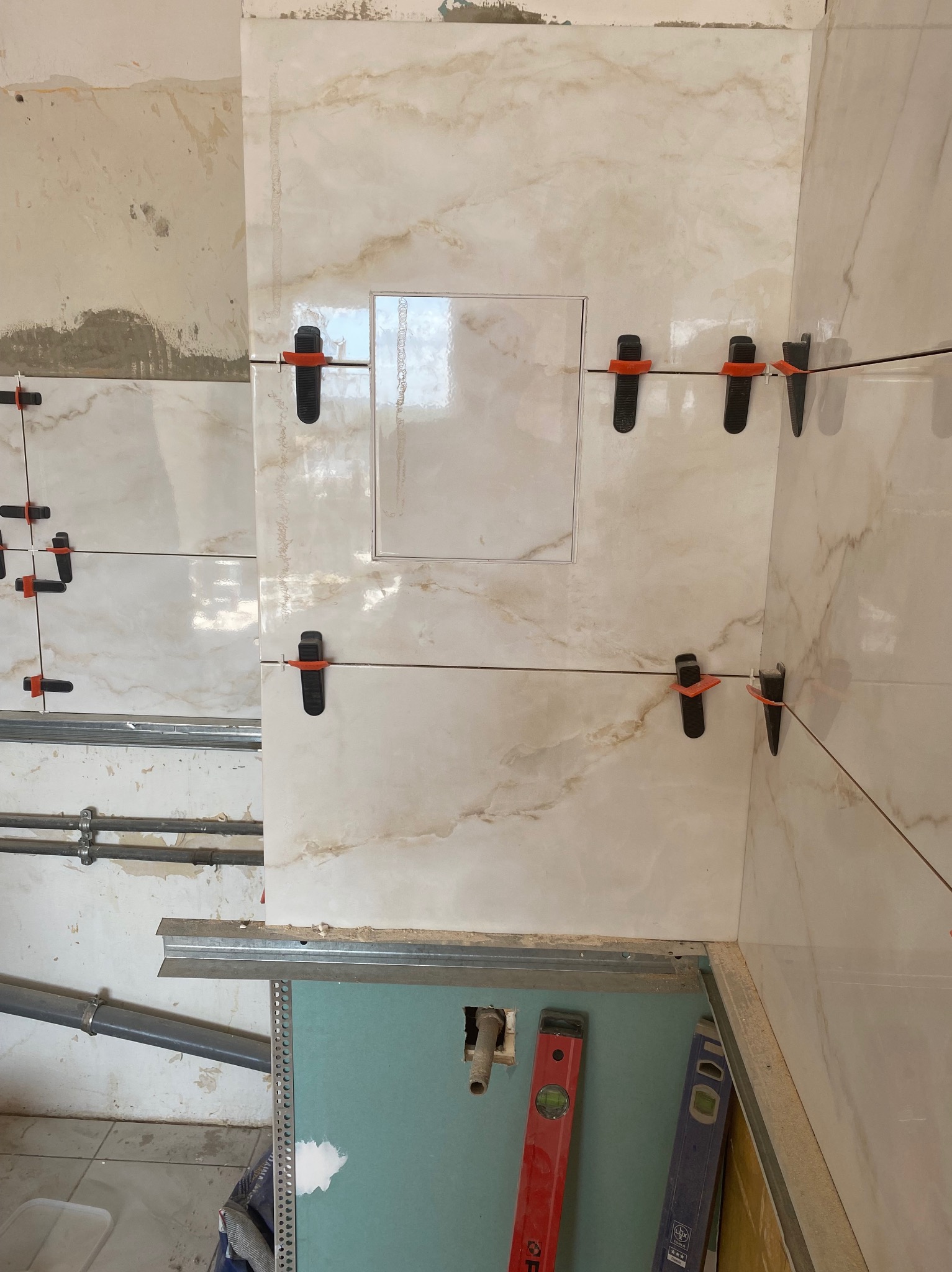 Układanie płytek imitujących marmur z użyciem klipsów poziomujących. Widoczny otwór rewizyjny, poziomice i fragment ściany przed wykończeniem.