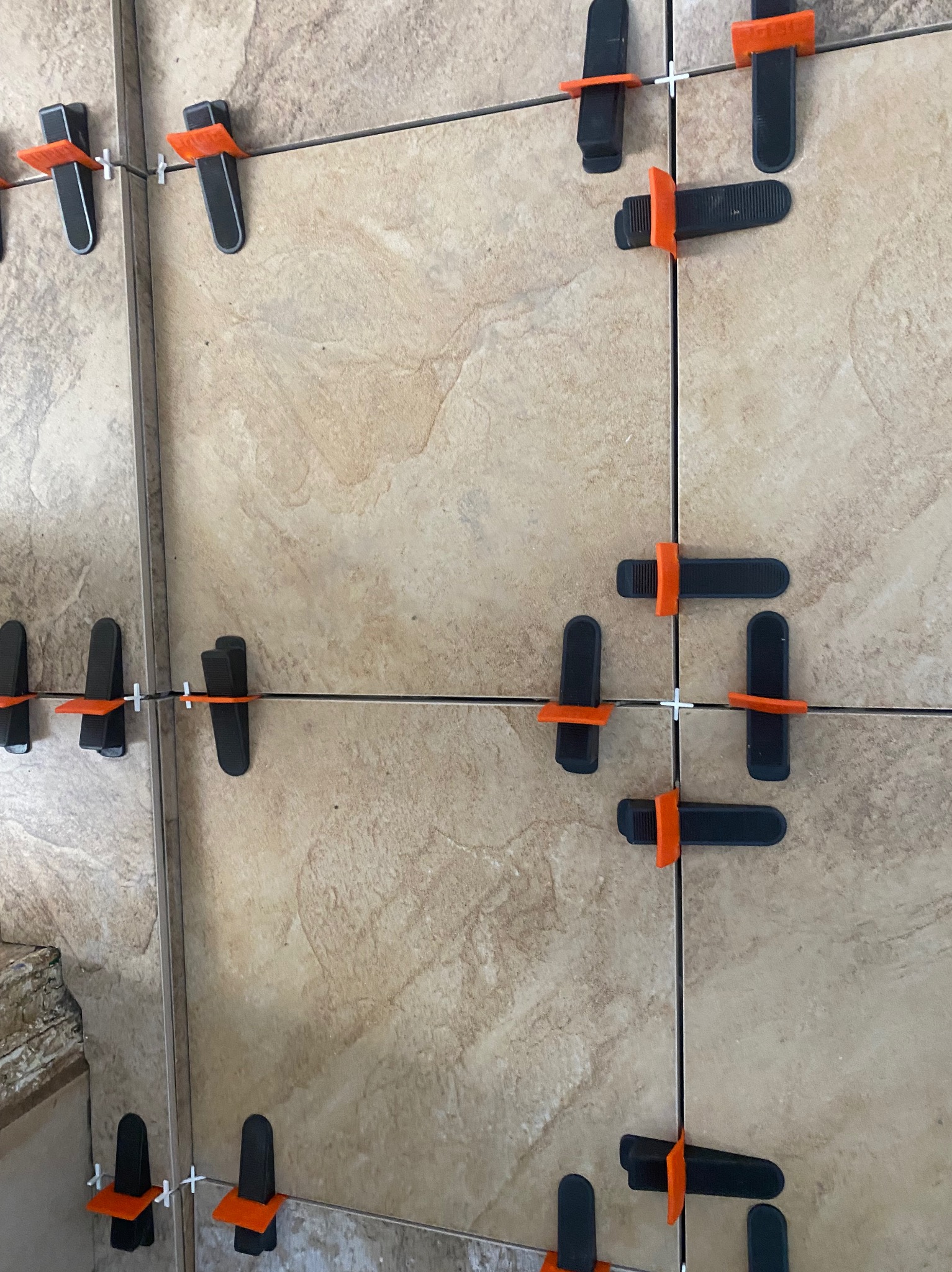 Ułożone płytki ceramiczne w beżowym odcieniu, z widocznymi klipsami poziomującymi w kolorze czarnym i pomarańczowym, oraz krzyżykami dystansowymi.