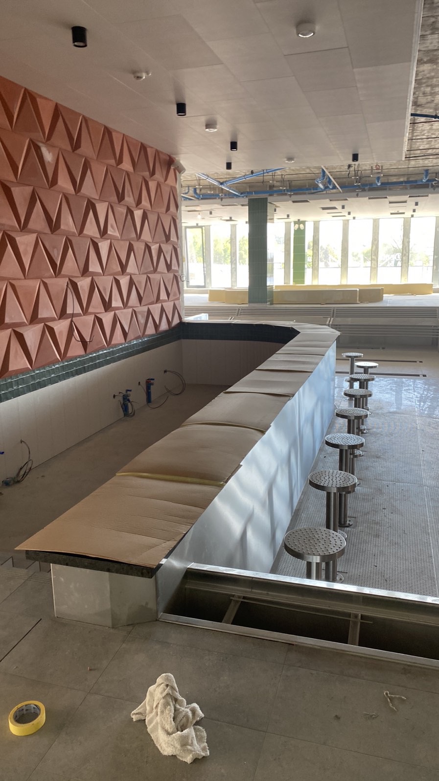 Wnętrze lokalu w Szczecinie w trakcie wykańczania. Widoczna ściana z trójkątną fakturą, bar i rząd metalowych stołków. Prace wykończeniowe w toku.