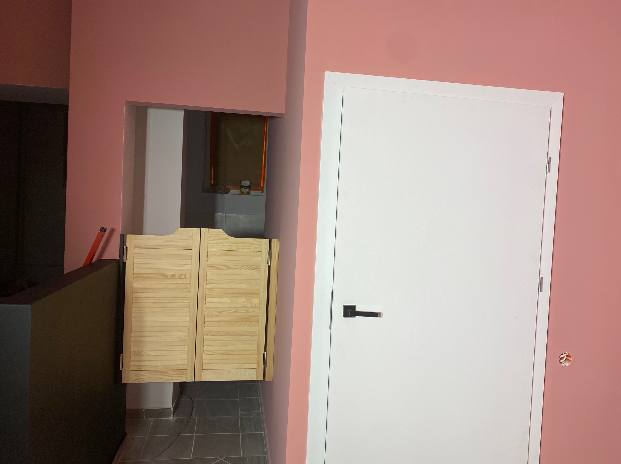 Różowa ściana z białymi drzwiami i drewnianymi drzwiami typu saloon. Widoczny fragment podłogi z szarych płytek. Detal wykończenia wnętrza.