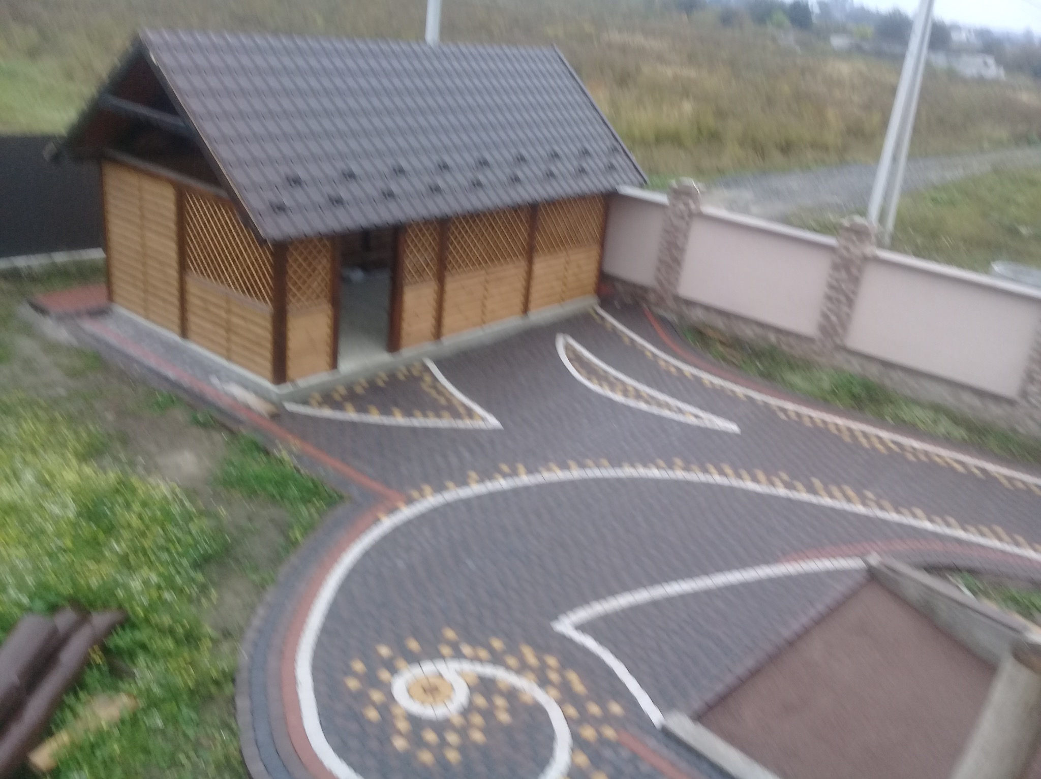 Altana z drewnianą kratownicą i brukowaną ścieżką z dekoracyjnymi wzorami, otoczona zielenią i niskim murem. Widok z góry.