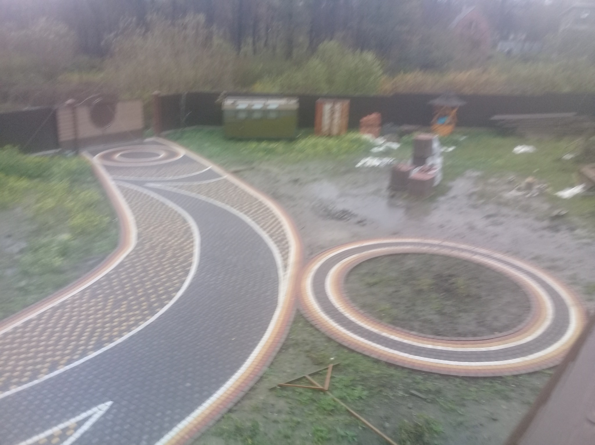 Kaskadowa ścieżka z kostki brukowej w ogrodzie, z elementami w kształcie okręgów. Widok z góry, w tle zieleń i ogrodzenie. Układanka z kostki w trakcie realizacji.