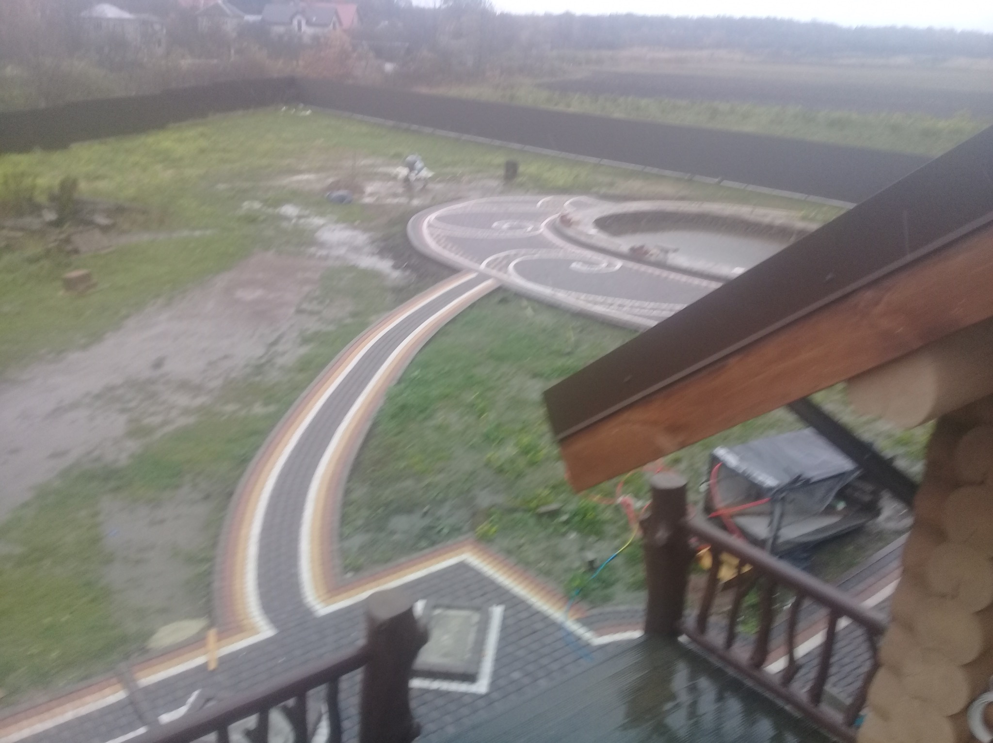 Widok z góry na ścieżkę z kostki brukowej w ogrodzie, z elementami dekoracyjnymi i oczkiem wodnym. Ujęcie z balkonu drewnianego domu w pochmurny dzień.